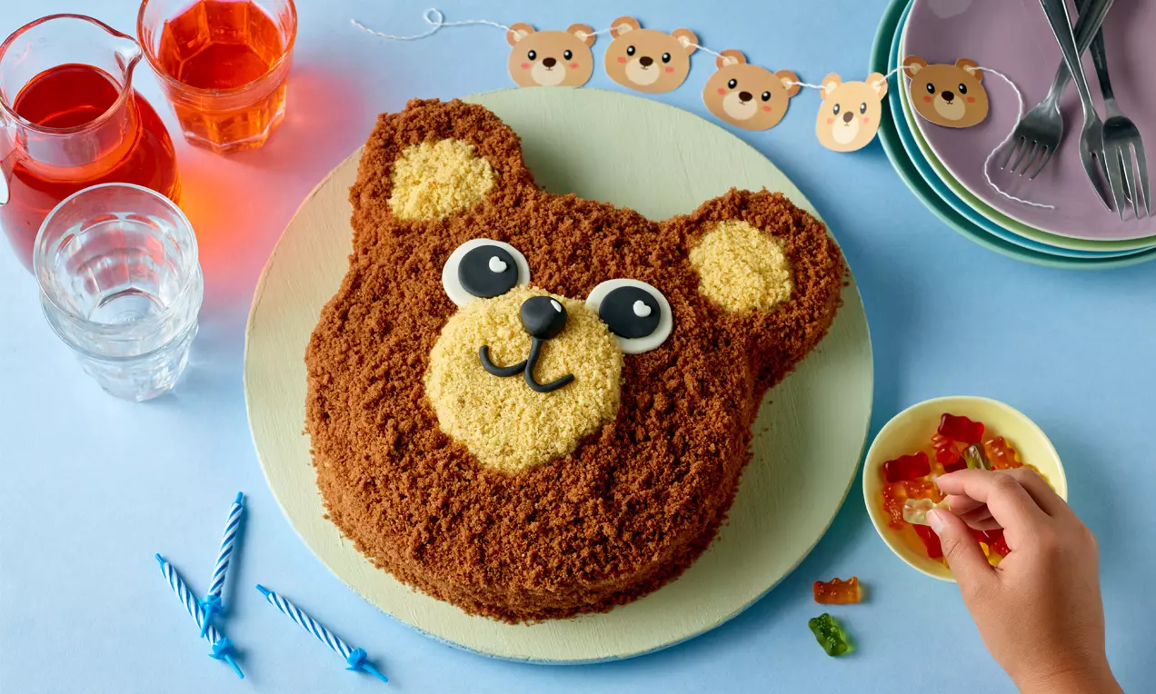 Braune Teddybär-Torte mit hellen Ohren und Schnauze, schwarzen Augen und Nase, auf hellem Teller neben bunten Dekorationen und Gummibärchen