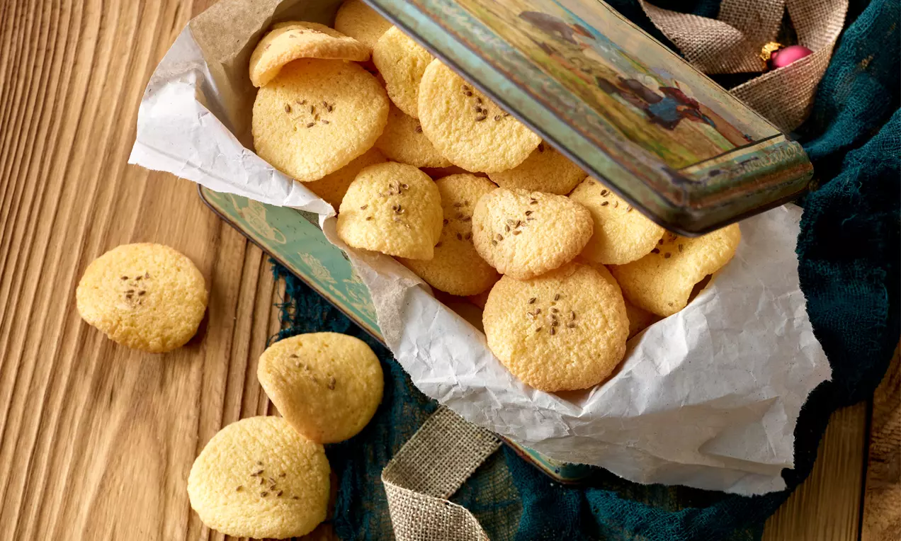Hellgoldene, leicht gebogene Anisbögen mit zarter Struktur und dunklen Anissamen, sorgfältig in einer nostalgischen dekorierten Blechdose auf weißem Papier gelegt