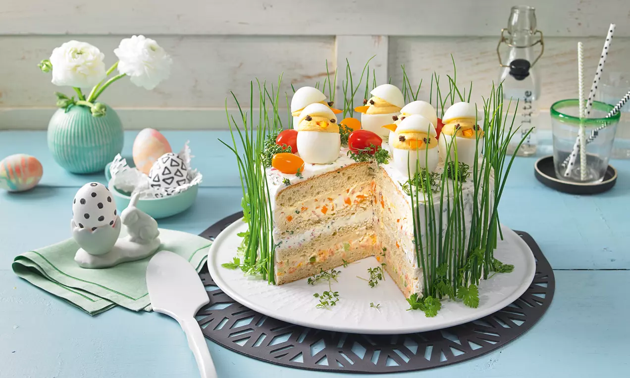 Pikante Sandwich-Torte in Schichten mit Creme, dekoriert mit Schnittlauch, Kresse und Ei-Küken auf weißer Platte mit schwarzer, runder Unterlage