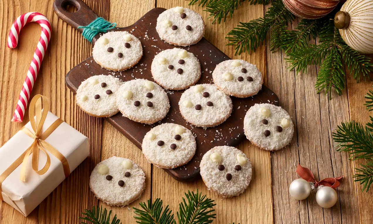 Runde Eisbär-Kekse mit weißer Kokosstreu, dunklen Schokodrops und cremefarbener Deko auf Holzbrett, umrahmt von Weihnachtsschmuck
