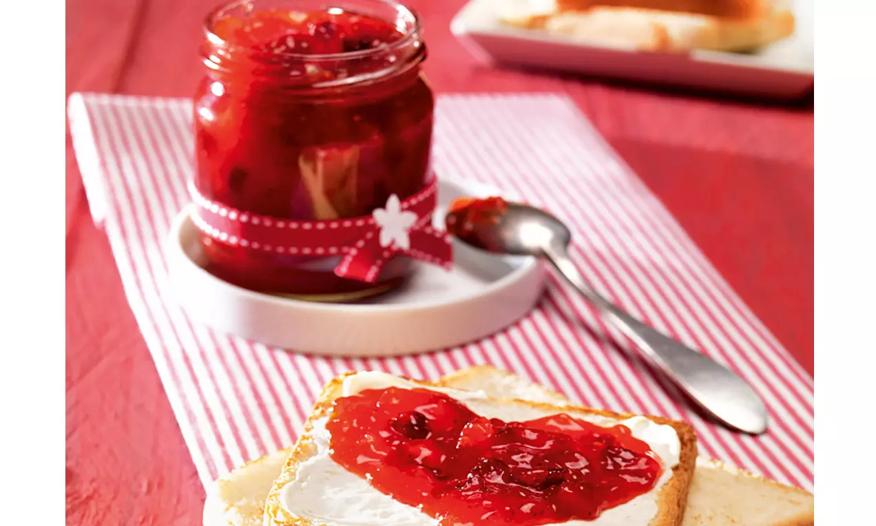 Glas mit tiefroter Erdbeer-Cranberry-Konfitüre auf weißer Unterlage, daneben Toastscheibe mit Konfitüre auf heller Creme und silbernem Löffel