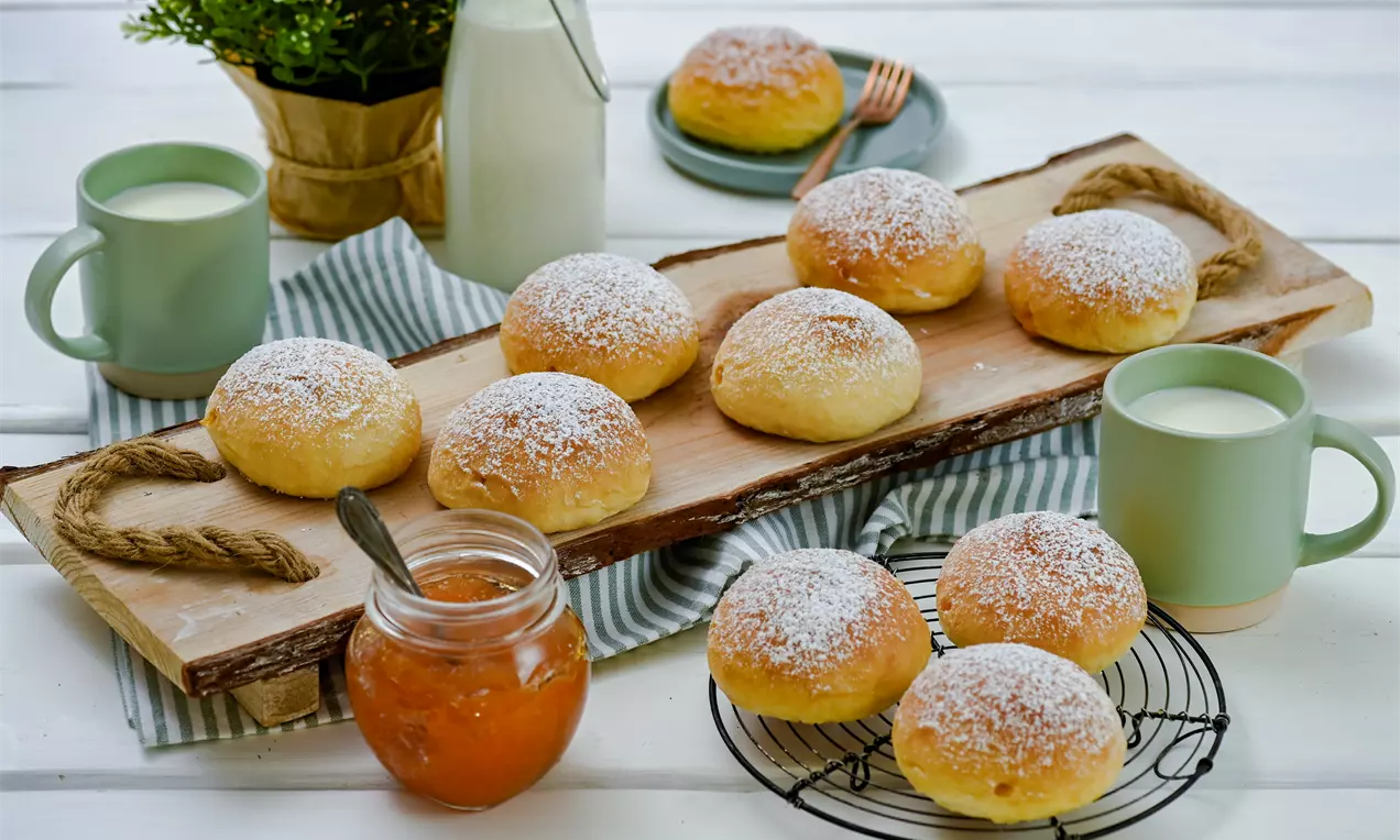 Goldgelbe Krapfen aus dem Backrohr, rund und flaumig, mit Zucker bestäubt, arrangiert auf Holzbrett und Gitterrost, neben Milch und Marmelade