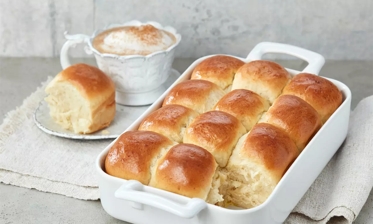 Goldbraune, glänzende Milchbrötchen in einer weißen Keramikform, ergänzt durch ein einzelnes Stück auf einem Teller neben einer Tasse Milchkaffee