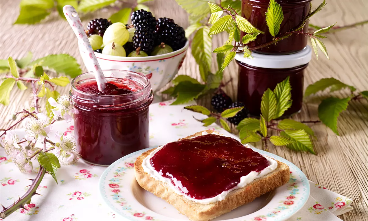 Samtige, dunkelrote Brombeer-Stachelbeer-Konfitüre in einem offenen Glas, angerichtet auf getoastetem Brot auf floralem Porzellanteller